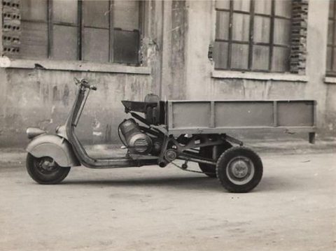 A sepia side view of a three-wheeler made from a scooter front with a wooden flat bed on two rear wheels. Opens a larger view