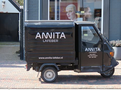Side view of a black three-wheeled van with white lettering, parked on brick paving in front of a shop window. Opens a larger view