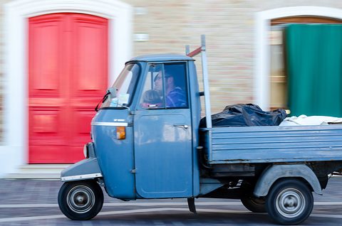Pale blue three-wheeled pick-up with a black bag in its bed, the background streaked by a panning shot past a red door. Opens a larger view