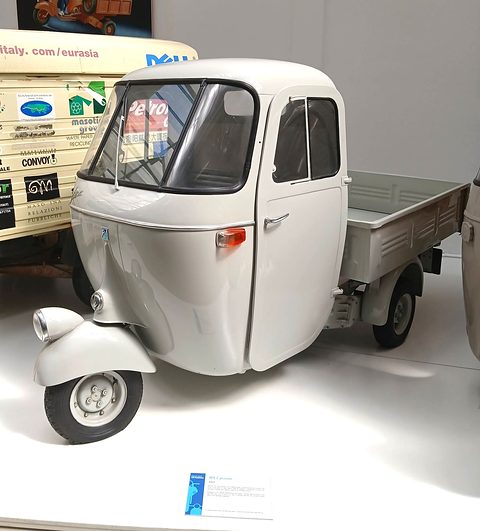 Pale grey three-wheeler with a rounded closed cab, a single front wheel under a scooter-style mudguard and a flat load bed.