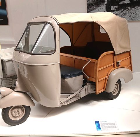 Pale grey three-wheeler with an open passenger body trimmed in wood, a dark bench seat and a beige folding canvas top.