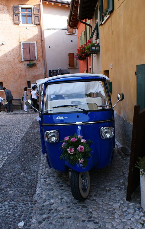 Blue three-wheeled passenger Ape with a white canopy and a bunch of pink roses on its nose, in a narrow cobbled lane. Opens a larger view