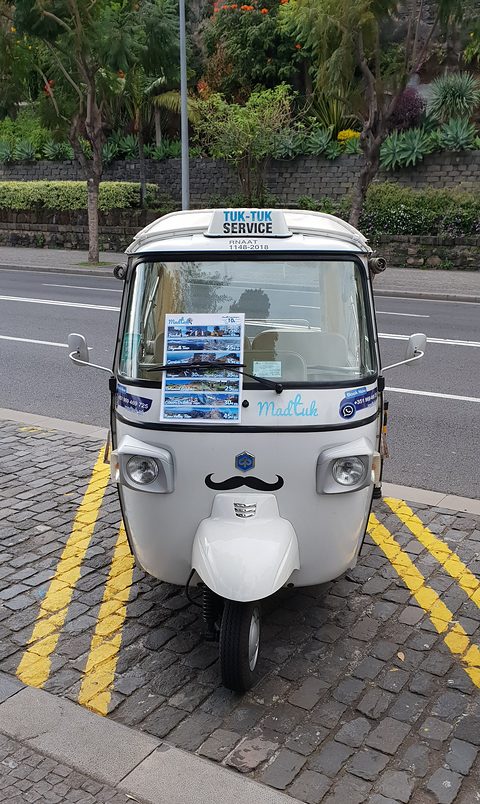 White three-wheeled passenger tuk-tuk head-on, a black moustache painted on its nose and a price board behind the windscreen. Opens a larger view