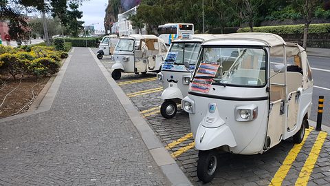 Four white three-wheeled tuk-tuks with white canopies parked in a line on cobbles beside a paved promenade. Opens a larger view