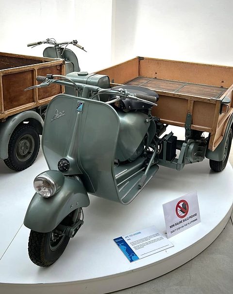 Grey-green Vespa-fronted three-wheeler with a wooden dropside box, on a white museum turntable. Opens a larger view