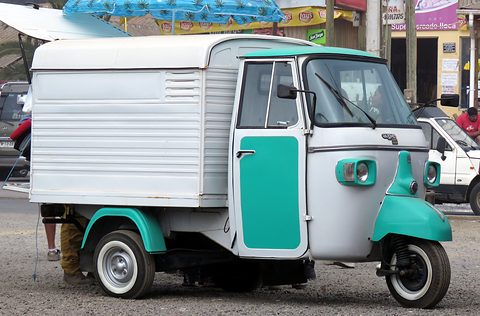 Three-wheeled van with a teal cab front and a ribbed white box body, parked under a market awning. Opens a larger view