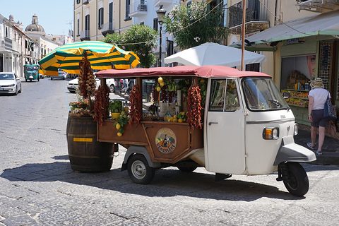 White three-wheeler with a wooden stall body hung with garlic and red chillies, a barrel of lemons beside it and a parasol above. Opens a larger view