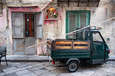 Dark green three-wheeler with wooden bed sides parked before a peeling pink and grey house front with a small shrine. Opens a larger view