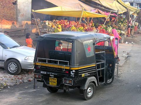Black three-wheeled taxi with yellow stripes and a rear rack, parked by a fruit market under yellow awnings. Opens a larger view
