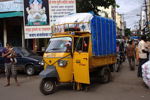 Yellow Indian three-wheeled truck with a blue tarpaulin load in a crowded street of shops and signs. Opens a larger view