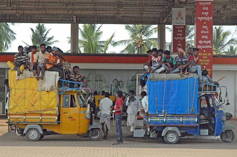 A yellow and a blue Indian three-wheeler under tarpaulins, young men sitting on their roofs, at a roadside shelter. Opens a larger view