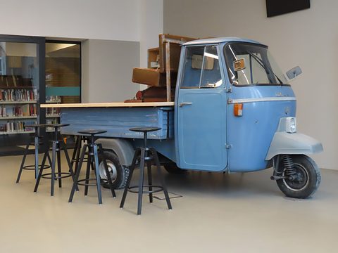 Pale blue three-wheeled pick-up indoors with a wooden counter top on its bed and tall black stools around it. Opens a larger view
