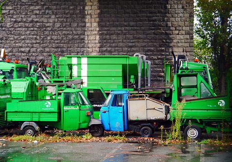 A row of green municipal three-wheeled utility trucks and one blue one, parked close together in front of a rusticated wall. Opens a larger view