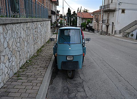 Rear of a small teal three-wheeler parked against a stone wall on a steep empty street lined with houses. Opens a larger view