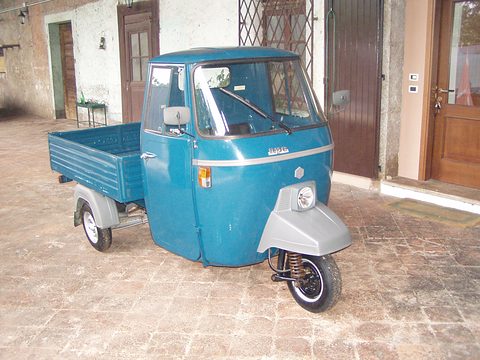 Teal three-wheeled pick-up with a grey front cowl, parked on a paved courtyard by old house doors. Opens a larger view