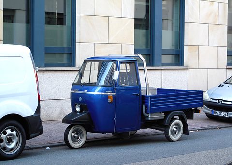 Side view of a shiny blue three-wheeled pick-up with whitewall tyres, between a white van and a silver car. Opens a larger view