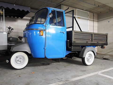 Side-on blue three-wheeled pick-up with a wooden-slatted bed and a mesh rear guard, under a striped awning. Opens a larger view