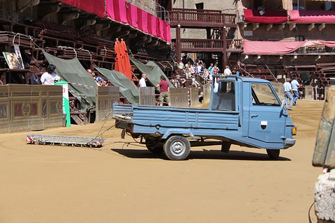 Blue three-wheeled pick-up towing a harrow over the sandy track of a great square, wooden grandstands and spectators behind.