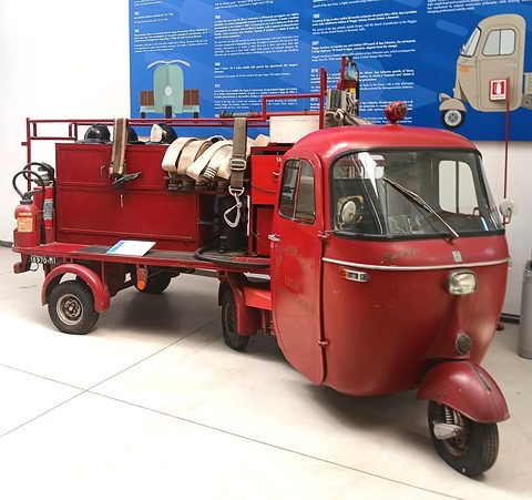 Red three-wheeled tractor cab coupled to a semi-trailer carrying hose reels and extinguishers, in a white museum room. Opens a larger view