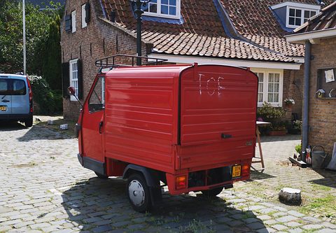 Red three-wheeled van with a tall boxy body and a roof rack, from behind, parked by brick cottages with tiled roofs. Opens a larger view