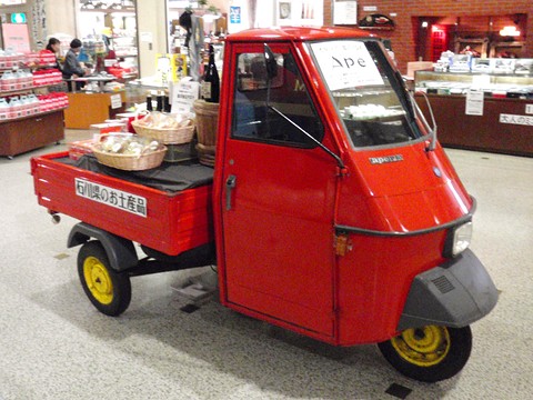 Red three-wheeled pick-up with yellow wheels carrying baskets of souvenirs, in a museum gift-shop hall. Opens a larger view