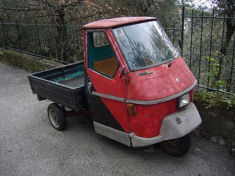 Faded red three-wheeled pick-up with a grey front cowl and a dark load bed beside a green railing and olive trees. Opens a larger view