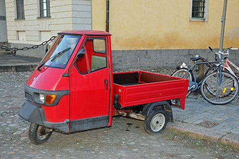 Bright red three-wheeled pick-up parked on cobbles beside a pair of bicycles and an ochre wall. Opens a larger view