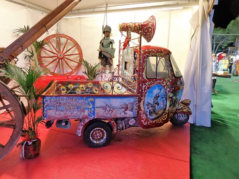 Three-wheeler covered in painted scenes and wrought-iron scrollwork, with a puppet knight and a red plume on top. Opens a larger view