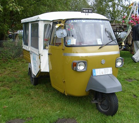 Olive and white three-wheeled taxi with a boxy white passenger body and a small roof sign, parked on grass. Opens a larger view