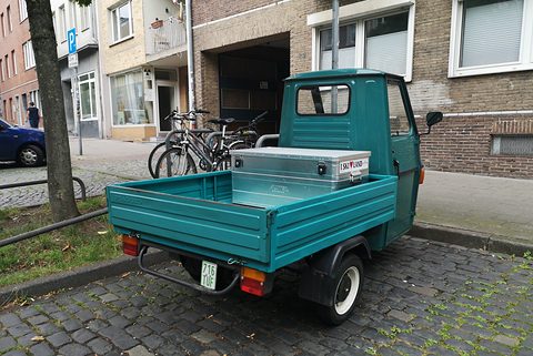 Rear quarter of a teal three-wheeler with an aluminium toolbox in its bed, parked on cobbles by bicycles and brick houses. Opens a larger view