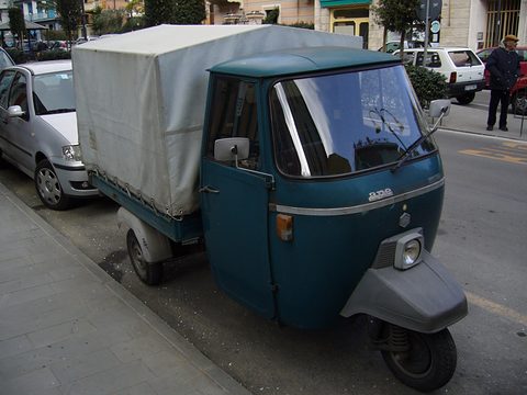 Dark teal three-wheeler with a grey canvas cover over the back, parked at a kerb beside a small silver car. Opens a larger view