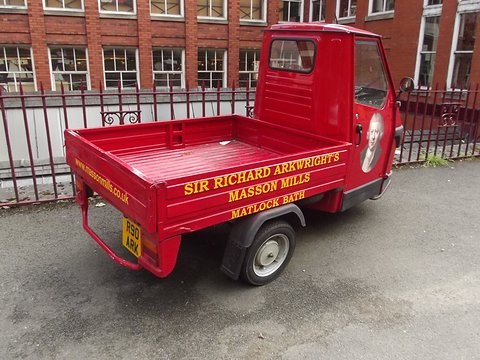 Red three-wheeled pick-up with yellow lettering on its dropside, parked in front of a brick mill with many windows. Opens a larger view