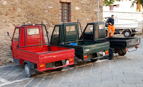 A red and two dark green three-wheeled pick-ups with wire-mesh rear window guards, parked nose to tail on a stone street. Opens a larger view