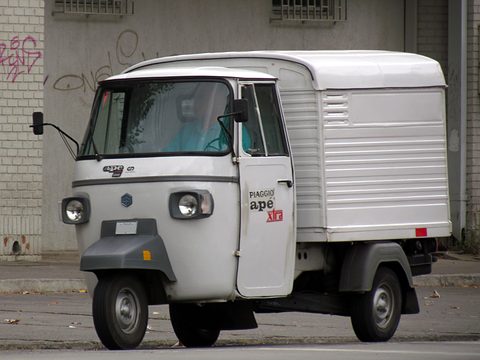 White three-wheeled van with an Ape Xtra decal on its door and a tall ribbed box body, against a graffitied wall. Opens a larger view