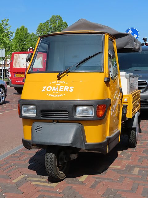 Head-on view of a yellow three-wheeler with ZOMERS on its nose and a canvas cover over its load, on brick paving. Opens a larger view