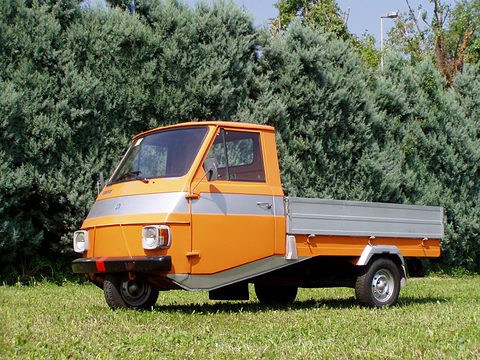 Orange three-wheeled pick-up with a grey band round its cab and a grey dropside bed, on grass before a hedge. Opens a larger view