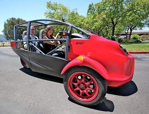 Three-wheeled two-seater with red front bodywork, a black tubular cage and red wheels, a man and a woman aboard.