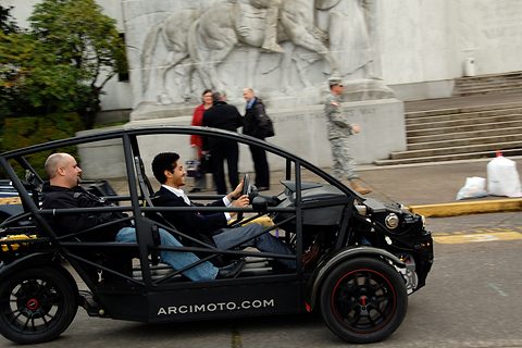 Black open-framed three-wheeler with ARCIMOTO.COM on its side and two men aboard in tandem, passing stone steps.