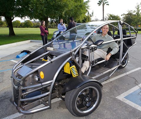 Three-wheeled tandem two-seater with a bare tubular steel frame and a yellow coil spring, a smiling driver in a car park. Opens a larger view