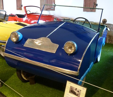 Dark blue rounded open two-seater with a polished nose plate and light blue lamp pods on artificial grass, a rope beside it. Opens a larger view