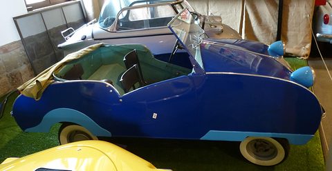 Long low dark blue open car in profile with whitewall tyres and a folded tan hood, a silver bubble car behind.