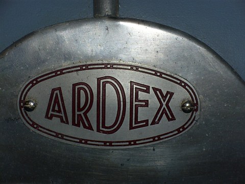 Oval metal badge with maroon outlined ARDEX lettering on a scratched polished nose panel.