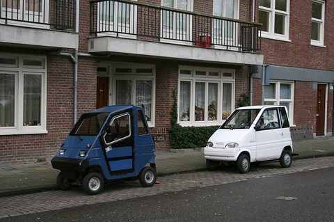 Small blue box-shaped two-door car and a small white car parked on a brick pavement in front of a terrace of flats.