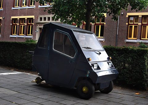 A tall boxy dark grey three-wheeler with a single front wheel, square headlamps and a large windscreen on a pavement. Opens a larger view