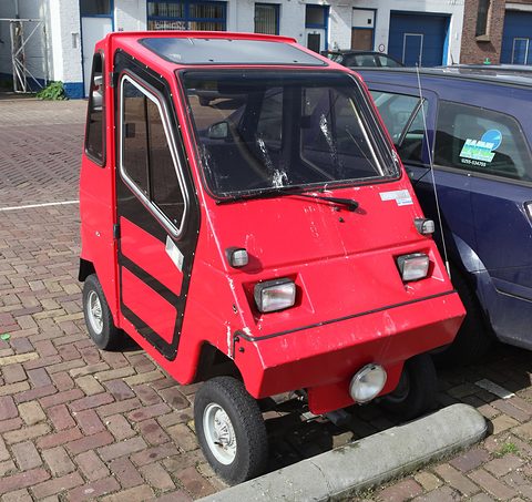 Red box-shaped two-door microcar with black door frames and twin square headlamps, parked beside a blue hatchback. Opens a larger view