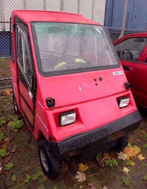 A boxy faded pink microcar with square headlamps and a black bumper, autumn leaves round its wheels. Opens a larger view