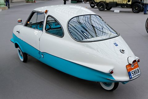 White and turquoise three-wheeler from the rear left, an orange number plate on its pointed tail, vintage cars beyond. Opens a larger view