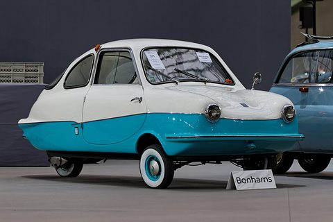 White and turquoise rounded coupé seen from the left side, whitewall tyres, sale cards in the windows, a blue Isetta behind.