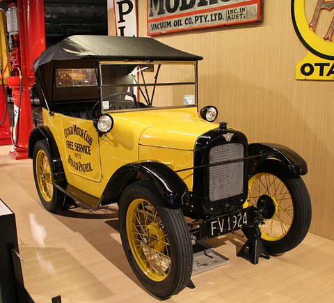 A yellow open tourer with black wings and lettering on its door before old fuel signs in a museum. Opens a larger view