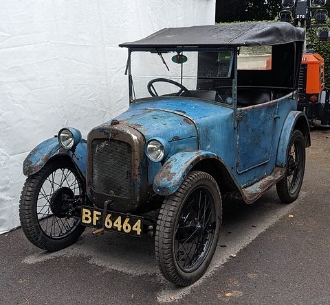 A small blue open tourer with worn, faded paint, a black hood and wire wheels against a white wall. Opens a larger view